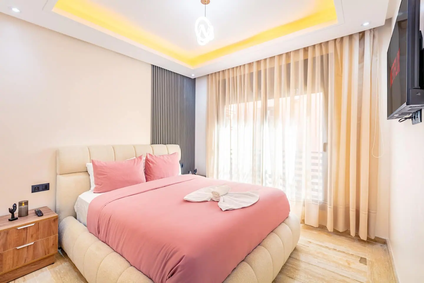 Chambre d'un appartement de Guéliz à Marrakech avec lit double à tête capitonnée, couette rose, serviettes pliées en éventail, rideaux clairs devant une baie vitrée et écran mural