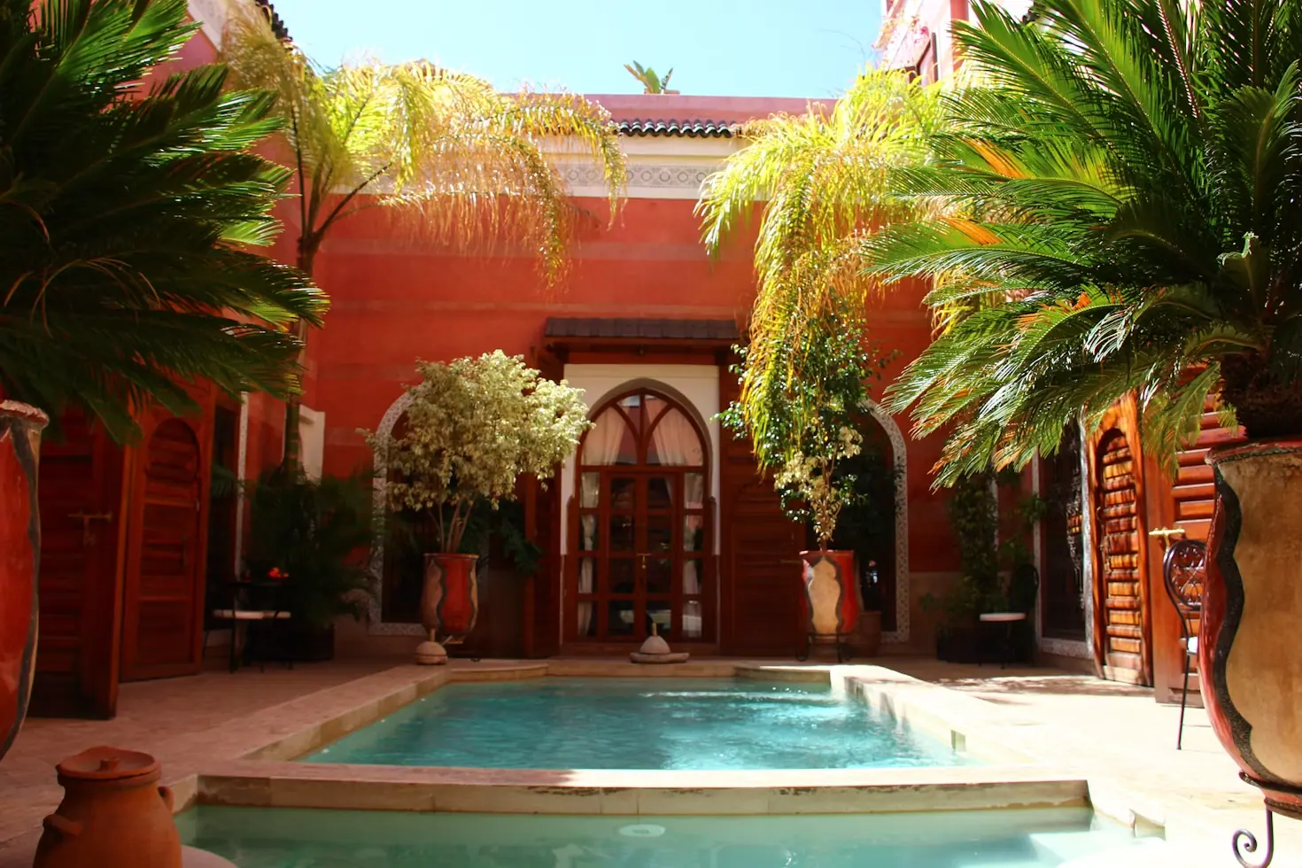 Patio d'un riad de la médina de Marrakech aux murs rouges, avec bassin, palmiers en pot et portes en bois sculpté