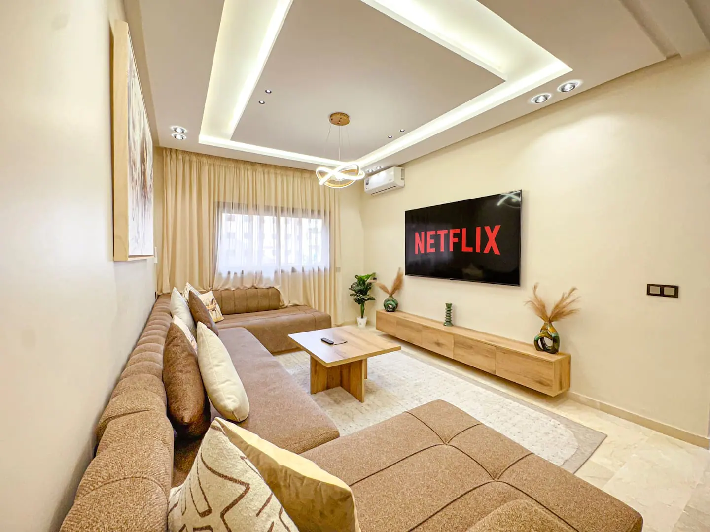 Salon d'un appartement marrakchi en tons beiges avec banquettes marocaines, faux plafond éclairé en corniche, grand écran mural, meuble bas en bois clair et climatiseur